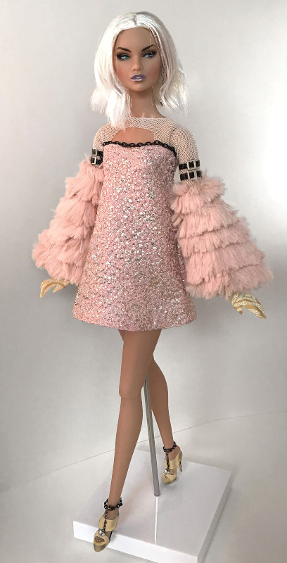 “She Owns the Night Too was created for an international Integrity Toys doll fashion design competition. Black chains edge the shimmery A-line bodice and custom shoes made in a pink and silver textured fabric. The mesh bodice with keyhole cutaway neckline has double-belted black straps that sit above layered blush-pink fur sleeves. Stylized gloves and drop ball earrings finish off the look.”