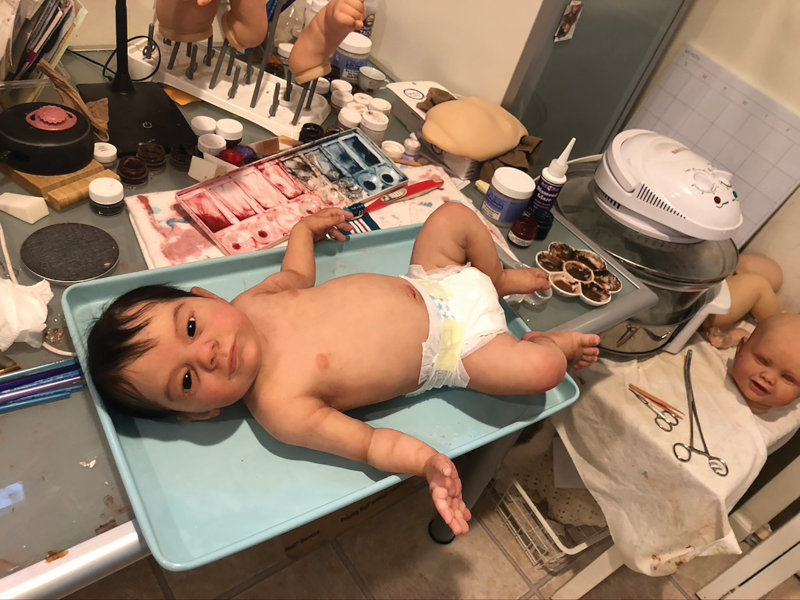 Evan, a lifelike baby doll, on a tray surrounded by paints and other dollmaking tools and materials.