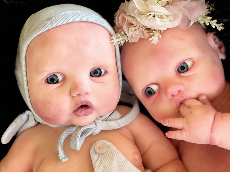 Two baby dolls lie next to each other, one with a flower headband and fingers in her mouth and the other with a blue head cap.